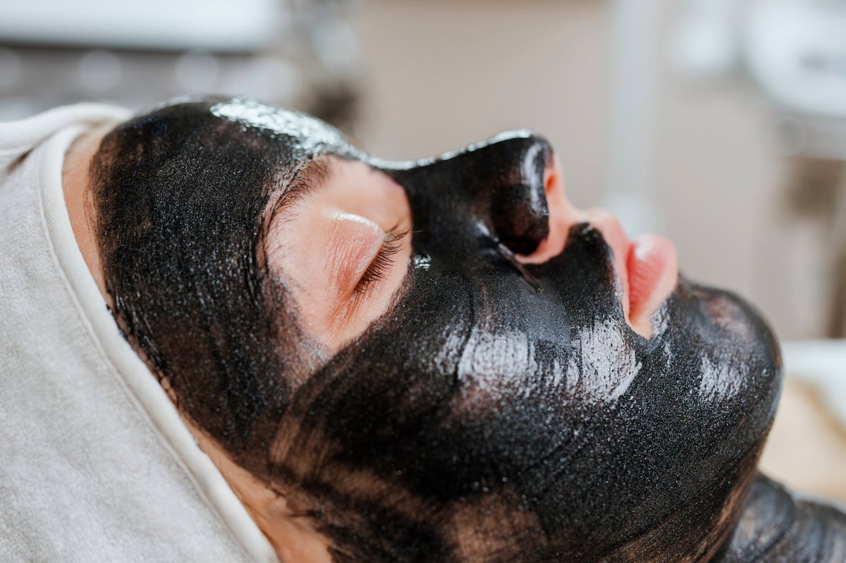 Cosmetologist applying a revitalizing black mask treatment to a young woman's face using a brush, enhancing her skin in a professional spa environment