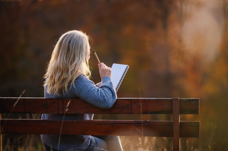 Thoughtful woman sitting on park bench and writing diary or inspiration notes during sunset